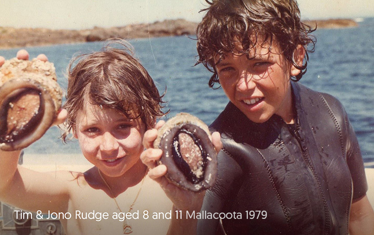 Yumbah founders Tim Rudge and Jono Rudge are pictured side by side as children. They are holding up an abalone each, while on a boat. White text on the bottom of the image reads: Tim & Jono Rudge, aged 8 and 11, Mallacoota 1979.
