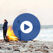 A blue play button is overlaid on an image of a woman on a beach with a bonfire behind her.