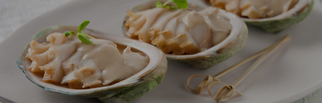 Three slow-cooked Abalone topped with a White Wine and Garlic Sauce, served in its shell on a white platter. Bamboo toothpicks to the side.