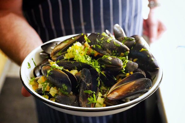 A plate of Smoked Mussels with Garlic, Butter and Herbs, held by a chef in a striped apron.